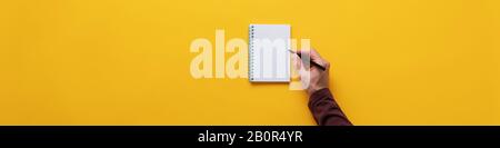 Wide view image of male hand holding black marker about to write in blank spiral notepad. Over yellow background. Stock Photo