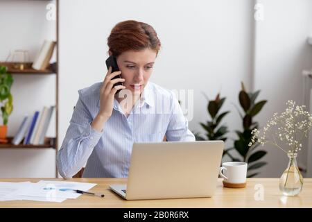 Busy businesswoman talking on phone, using laptop, looking at screen, reading information or email, employee consulting client, solving problem or dis Stock Photo