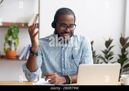 Smiling African American employee in headphones using laptop, looking at screen, making video call or watching webinar, writing notes, distance learni Stock Photo