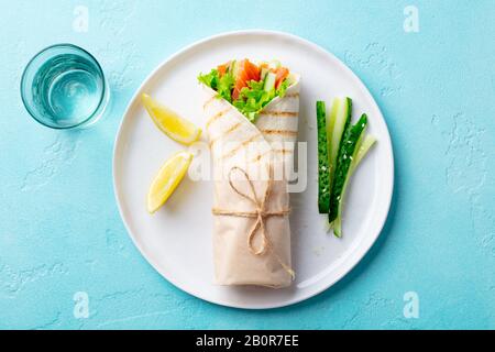 Wrap sandwich, roll with salmon, cucumber, salad on a white plate. Blue background. Top view. Stock Photo