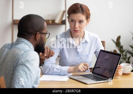 Diverse colleagues analyzing diagrams together, using laptop, looking at screen, working on project, Caucasian businesswoman, mentor explaining busine Stock Photo