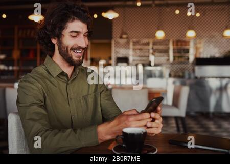Cheerful handsome young man relaxing in modern cafe using cellphone while drinking coffee Stock Photo