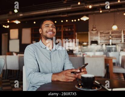 Handsome young african man laughing while reading funny message on phone sitting in modern cafeteria during free time Stock Photo