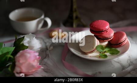 Close up view of Colourful Pastel Macarons on white ceramic plate with tea cup, pink ribbon and flower decorated on marble desk Stock Photo