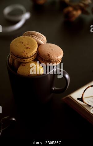 Cropped shot of chocolate macarons in black coffee cup next a book and eye glasses in workspace Stock Photo