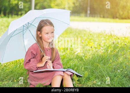 child reading a book. little girl under an umbrella sits on green grass on a bright sunny day Stock Photo
