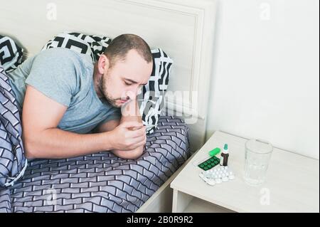 A man with a cold is lying in bed and taking his temperature. Stock Photo