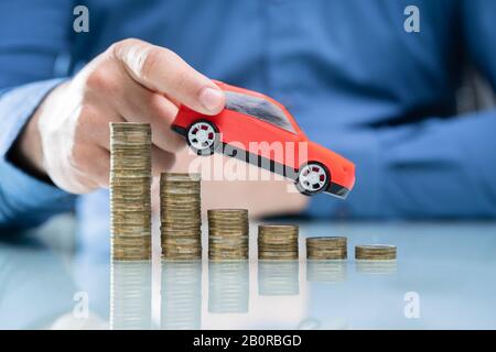 Businesswoman's Hand Driving Blue Car On Declining Stacked Coins Over Desk Stock Photo