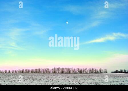 Full moon over plowed field covered with snow, line of trees on the hills on horizon, winter landscape, bright blue cloudy sky Stock Photo