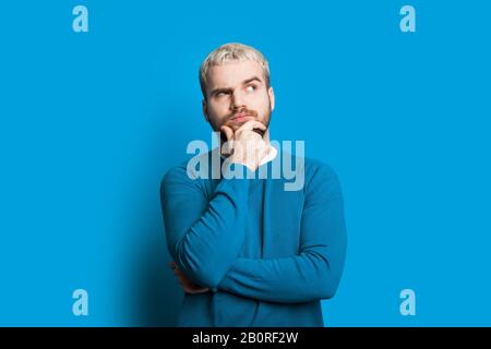 Blonde caucasian youngster touching his chin and looking aside while posing on a blue background in a sweater Stock Photo