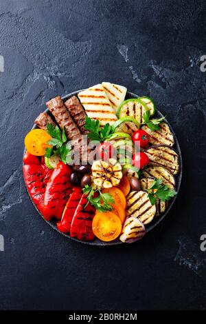 Grilled meat kebabs and vegetables on a black plate. Black stone background. Top view. Stock Photo