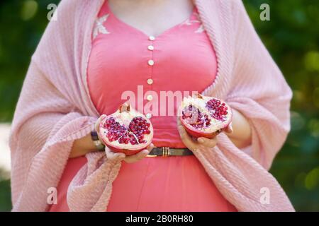 A curvy woman in pink dress and cardigan is holding in her hands two pieces of pomegranate Stock Photo