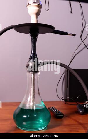 Black matte hookah with clear water with a black hose on the table next to the remote control in the lounge bar on a light background. Hookah cafe lei Stock Photo
