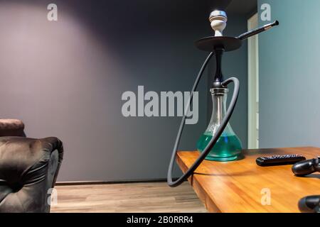Black matte hookah with clear water with a black hose on the table next to the remote control in the lounge bar on a light background. Hookah cafe lei Stock Photo