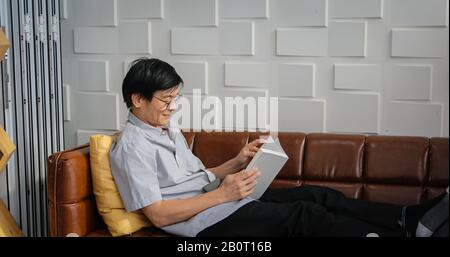 Senior Asian man reading book on sofa in living room at home ,Portrait of Asian elderly man is Relaxing and Happiness With Read a Magazine Stock Photo