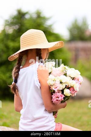 Girl with a bouquet of white and pink peonies. Sunny summer day. Girl in a hat with wide brim. Back view. Girl with two braids. Stock Photo