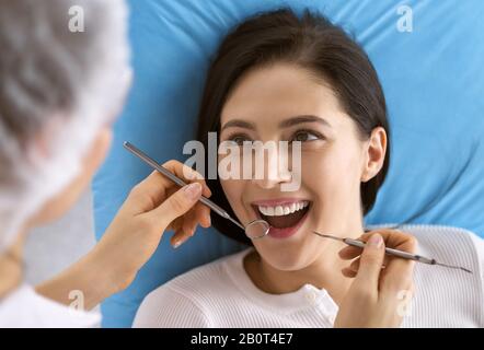 Smiling brunette woman being examined by dentist at dental clinic. Hands of a doctor holding dental instruments near patient's mouth. Healthy teeth Stock Photo