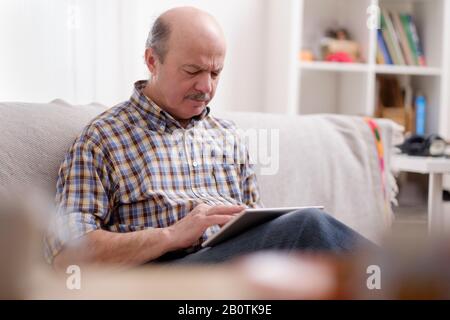 Handsome seniorman using a tablet at home sitting on a sofa in his living room browsing the internet with a smile Stock Photo