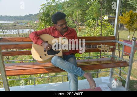 Close up of a person playing guitar sitting on a bench in a park or garden in west Bengal, India, selective focusing Stock Photo