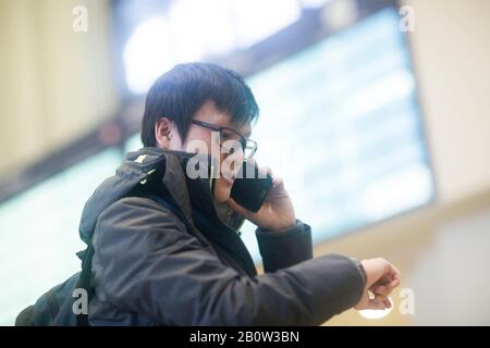 Young Asian man wearing glasses standing in railway station foyer, checking his watch and using mobile phone. Stock Photo