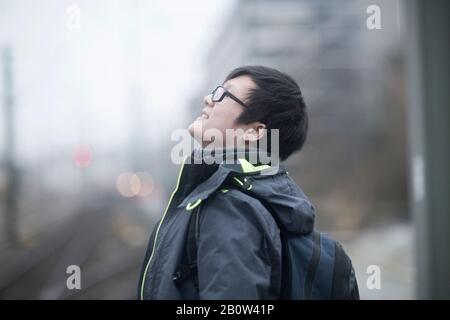 Young Asian man wearing glasses standing on railway platform, looking up, waiting for train. Stock Photo