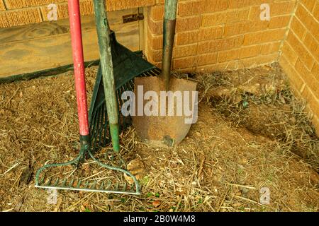 A garden rake, leaf rake, hoe and rusty shovel leaning against a brick wall. Stock Photo