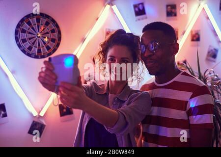 Young smiling woman holding camera and making a selfie portrait with young African man at a party Stock Photo