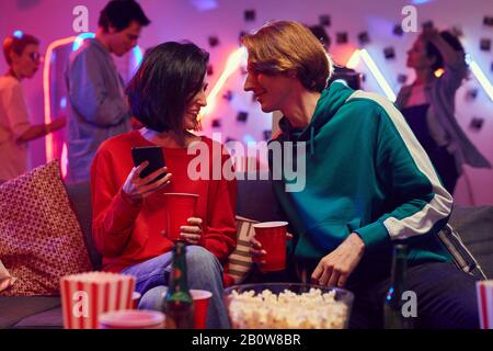 Young man sitting on sofa and talking to the young woman while she using her mobile phone they are at the party Stock Photo