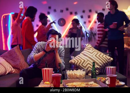Young man sitting on sofa and drinking alcohol while his friends dancing in the background during a party Stock Photo