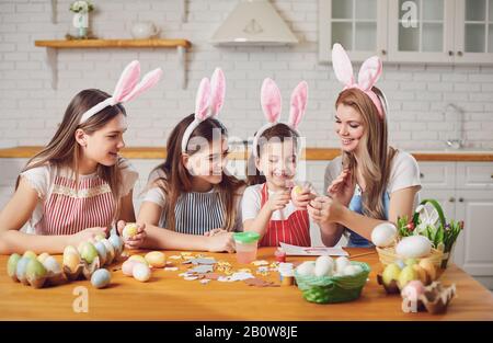 Happy easter. Mother and daughters with rabbit ears decorate Easter eggs at a table in kitchen. Stock Photo