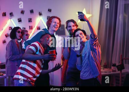 Group of happy friends standing and posing at camera of mobile phone for selfie portrait at party Stock Photo
