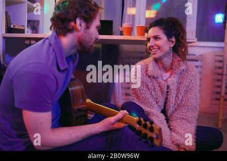 Young happy woman sitting on the floor and smiling while young man sitting near by her and playing on guitar at home Stock Photo