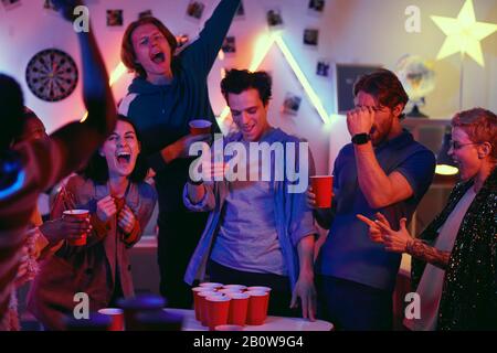 Excited young people drinking beer and playing in beer pong during home party Stock Photo