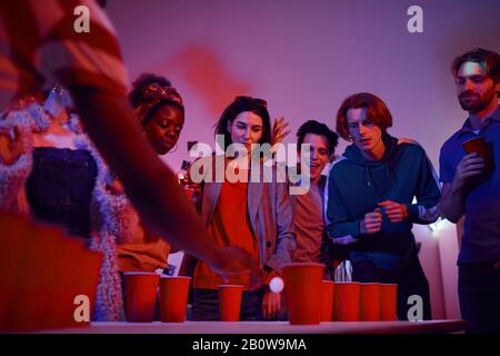 Group of teenagers standing around the table with glasses of beer and playing in beer pong at party Stock Photo