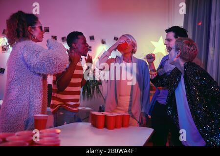 Mature woman standing and drinking beer while playing in beer pong with friends at the party Stock Photo