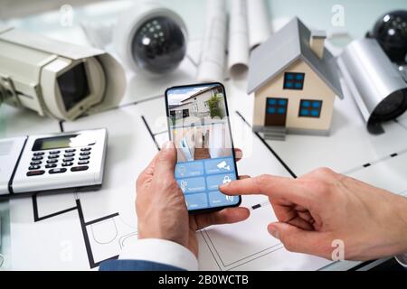 Person Hand Using Home Security System On Mobilephone With On Blueprint With Security Equipment Stock Photo