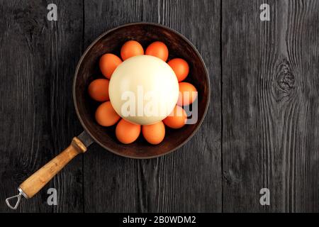Ostrich egg surrounded by chicken eggs in an old cast-iron skillet, which is standing on an old black wooden surface, top view, copy space.