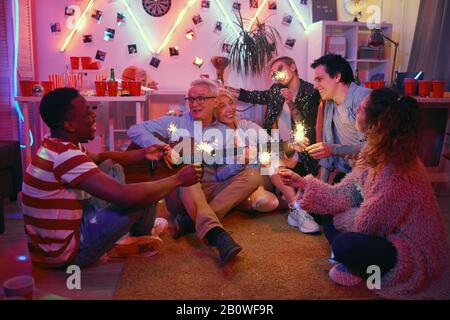 Senior couple sitting on the floor and playing guitar among other people who listening to them at home party Stock Photo