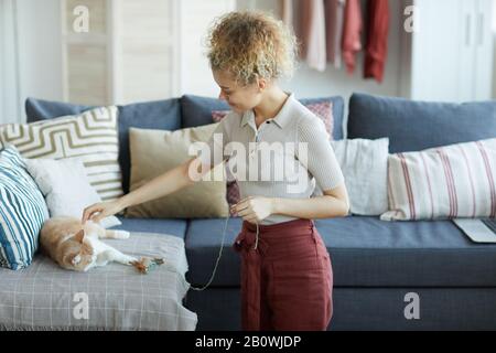 Young woman with curly hair holding toy of her cat and playing with her cat on the sofa at home Stock Photo