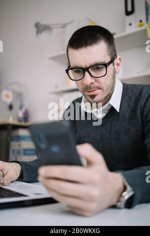 https://l450v.alamy.com/450v/2b0wkxg/young-man-with-glasses-looking-at-his-phone-businessman-wearing-glasses-looking-at-his-out-of-focus-phone-while-working-in-the-office-2b0wkxg.jpg