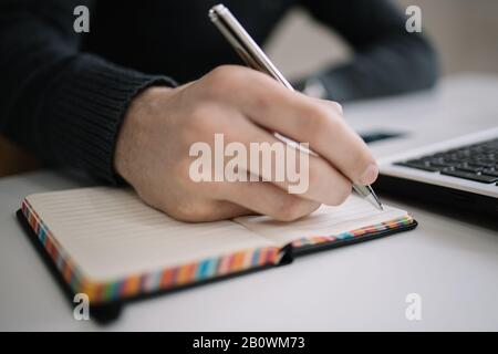 Close-up view of male hand writing in notebook. Man taking notes and writing in notepad with pen on desk. Stock Photo