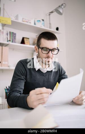 Male architect checking project on paper sheets. Young focused employee wearing glasses working with documents in office. Stock Photo