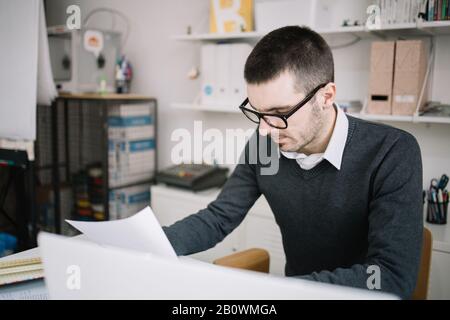 Office worker reading paperwork in the office. Focused employee wearing glasses and reading documents in coworking space. Stock Photo