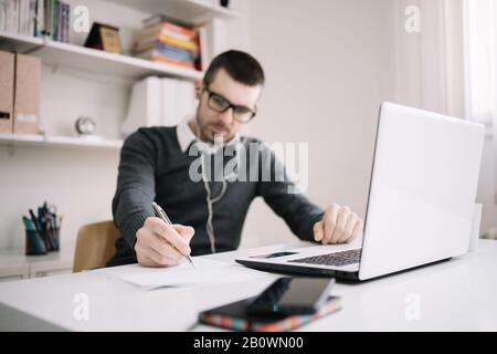 Man wearing headphones writing with pen at paper on work desk. Focused man having an online meeting while sitting at desk and taking notes in notepad. Stock Photo