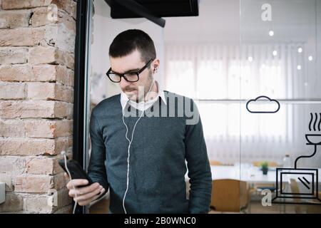 Slim man wearing headphones and glasses looking phone. Bearded guy with headphones holding smartphone while standing in the office. Stock Photo