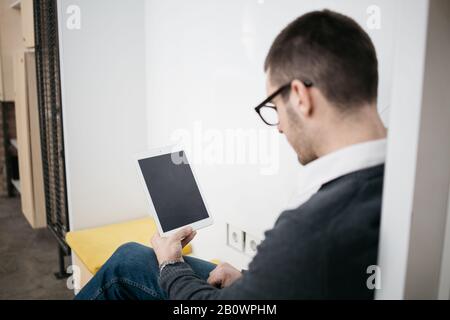 Man with glasses holding tablet with blank screen. View from back of an employee working on tablet with clean desktop at office. Stock Photo