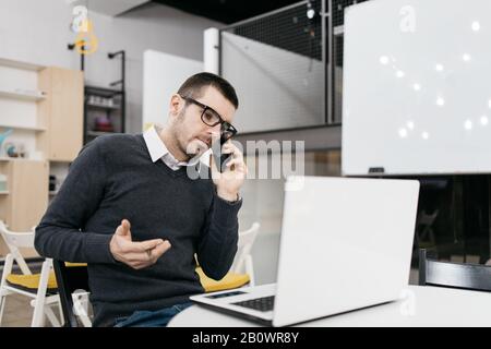 Man with glasses using phone and making gestures. Office worker using laptop while talking on phone and doing hand gestures. Stock Photo