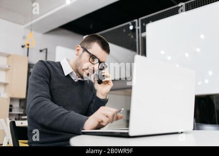 Male worker talking on phone and looking at laptop. Serious man with glasses speaking on phone and holding a pen while looking at laptop. Stock Photo