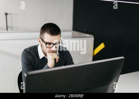 Entrepreneur reading online articles and magazines at office desk. Male employee wearing glasses working on a computer and touching his chin in the of Stock Photo