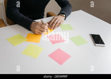 Hand holding pen writing on blank colorful sticky note. Cropped man writing on blank colored sticky notes on desk. Stock Photo